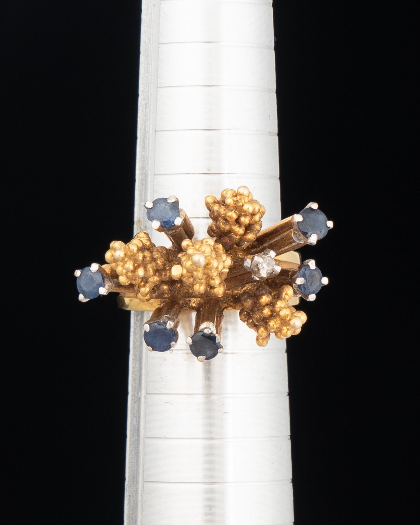 Gold ring with blue gemstones on a white stand against a black background